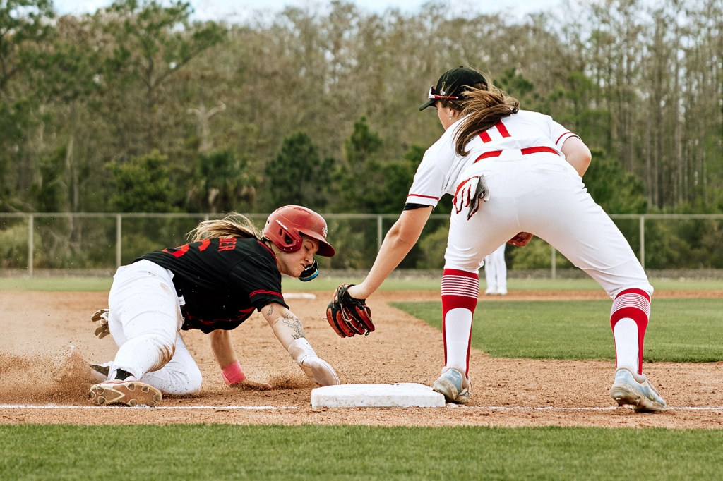 A baseball player slides toward a base as another player reaches down with her glove.