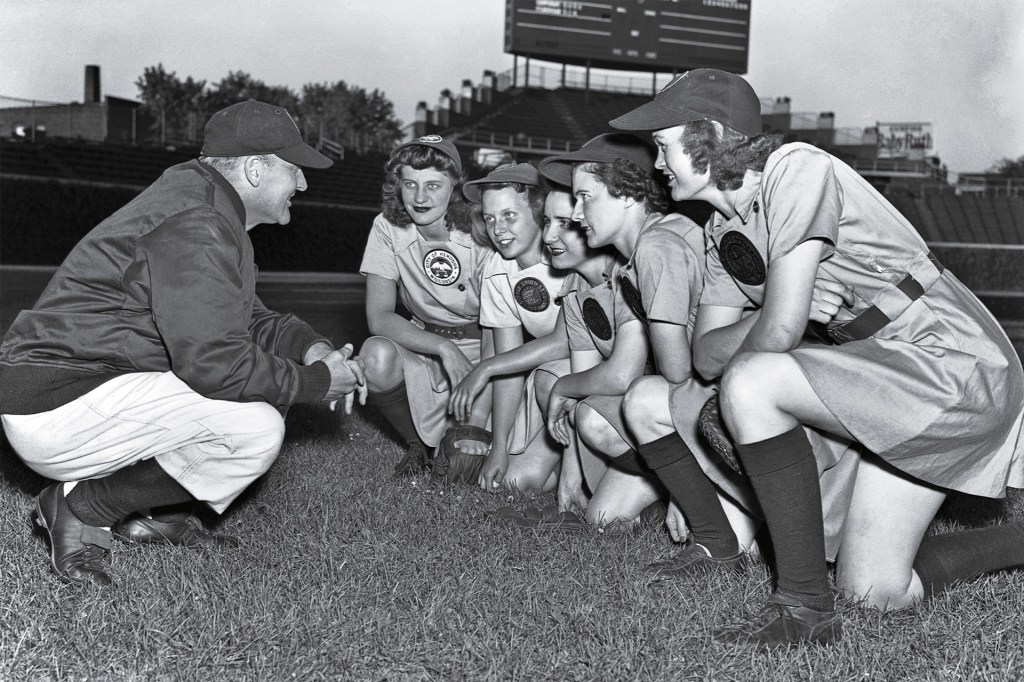 In an old black-and-white photo, a baseball coach talks with a group of women players on the field.