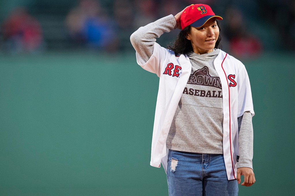 A young woman in a baseball jersey and red cap stands on a baseball field.