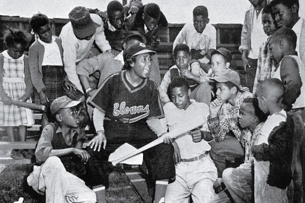In a black-and-white photo, a woman baseball player sits with a group of children gathered around her.