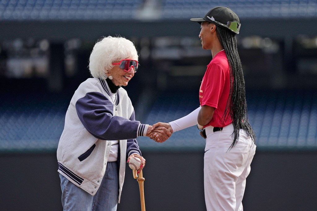 An older woman holding a baseball and cane shakes hands with a baseball player on the field.