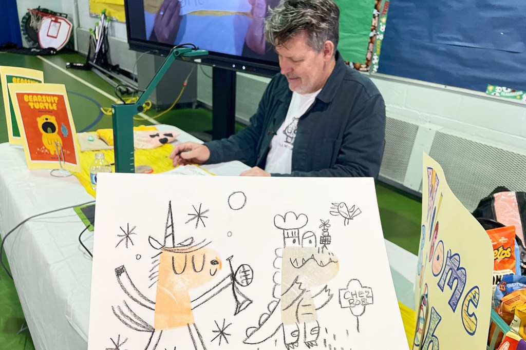 An author sits at a table, a drawing of two funny characters in the foreground.