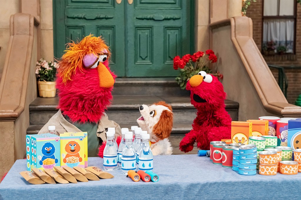 Two red puppet characters and a small dog puppet behind a table filled with food and supplies.