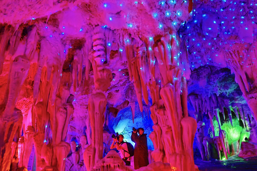 People explore a cave filled with colorful lights and hanging ice formations.