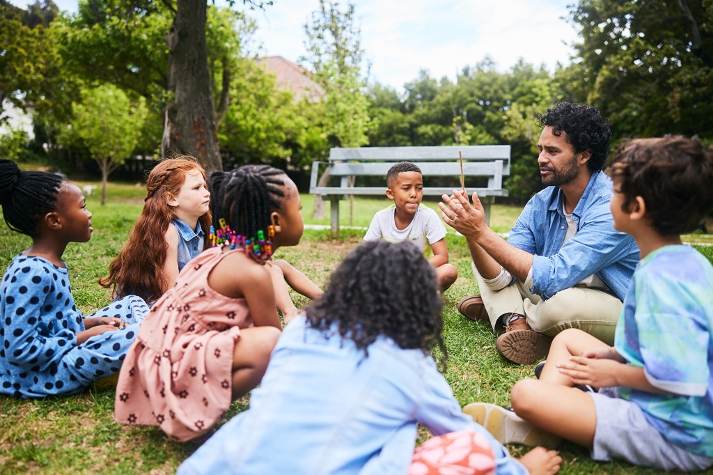 A teacher and a group of children sit in a circle on the grass during an outdoor lesson.