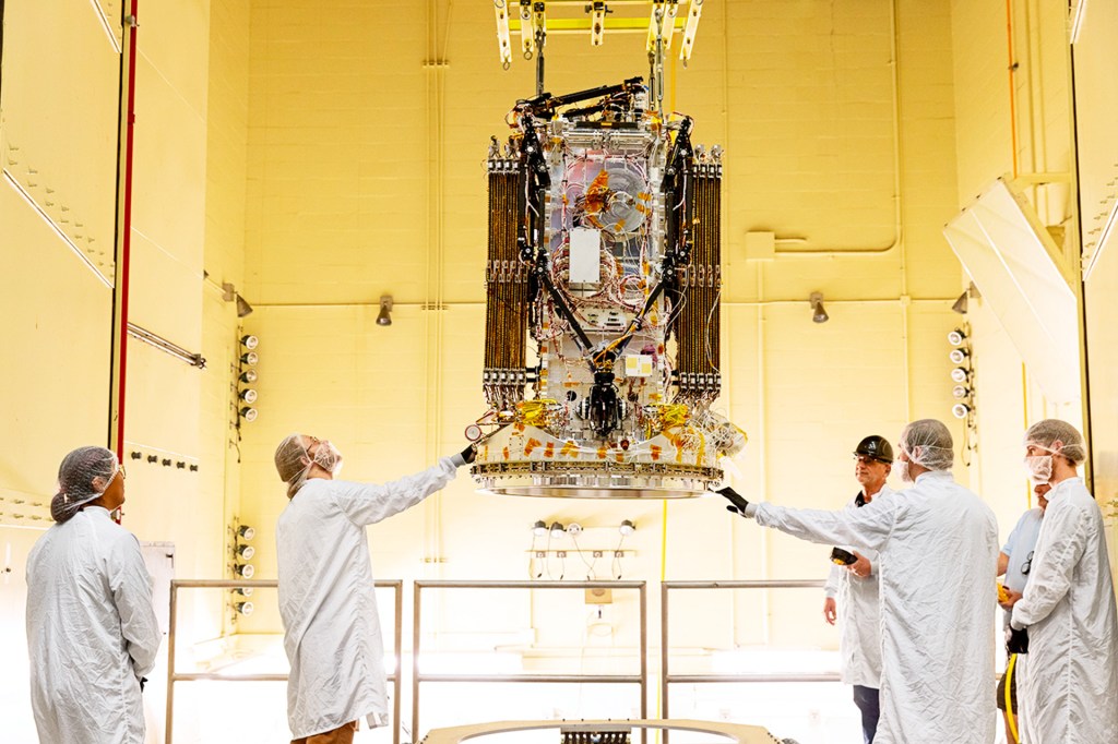 Workers in protective clothing examine a large spacecraft inside a bright room.
