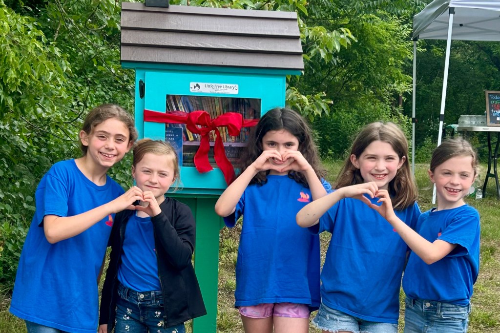 Five girls in matching blue shirts make heart shapes with their hands in front of a small outdoor library.