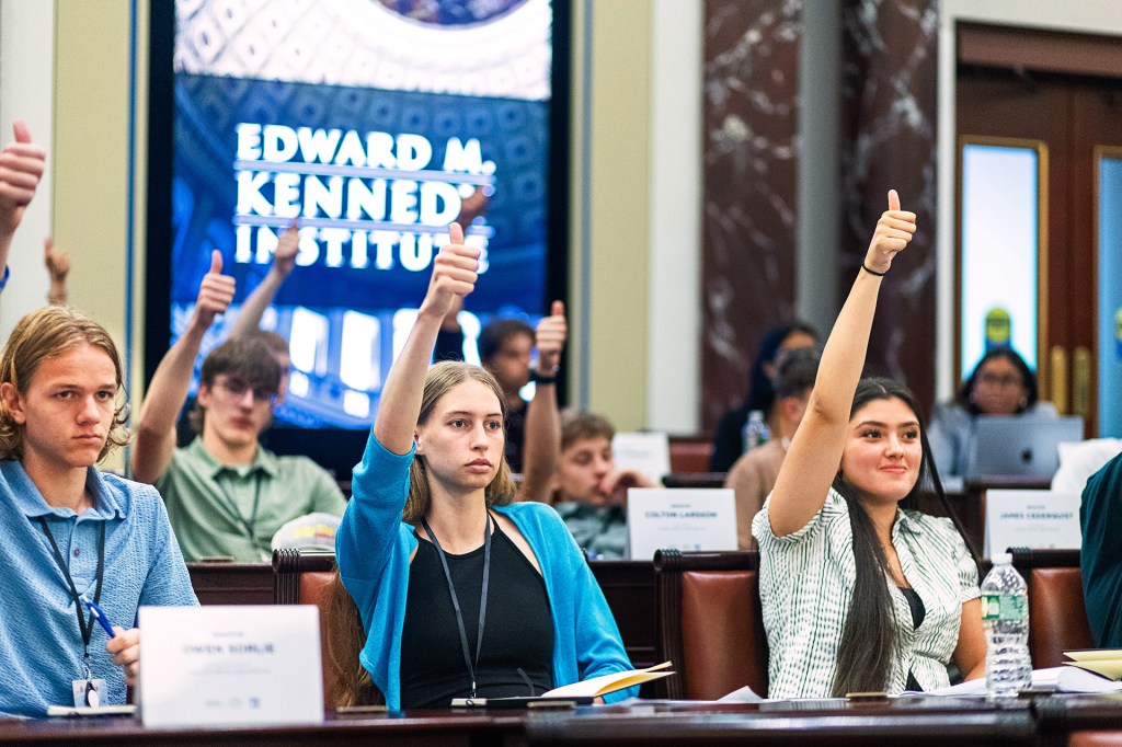 Students seated at tables raise their hands and give thumbs-up signs during a group meeting.