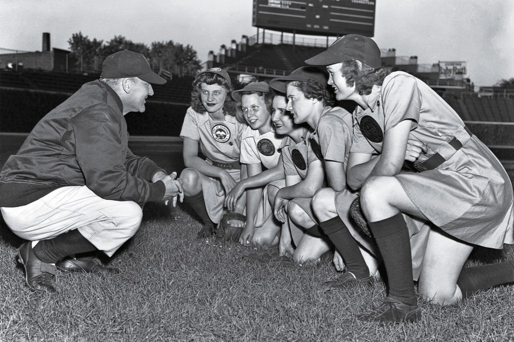 In an old black-and-white photo, a baseball coach talks with a group of women players on the field.