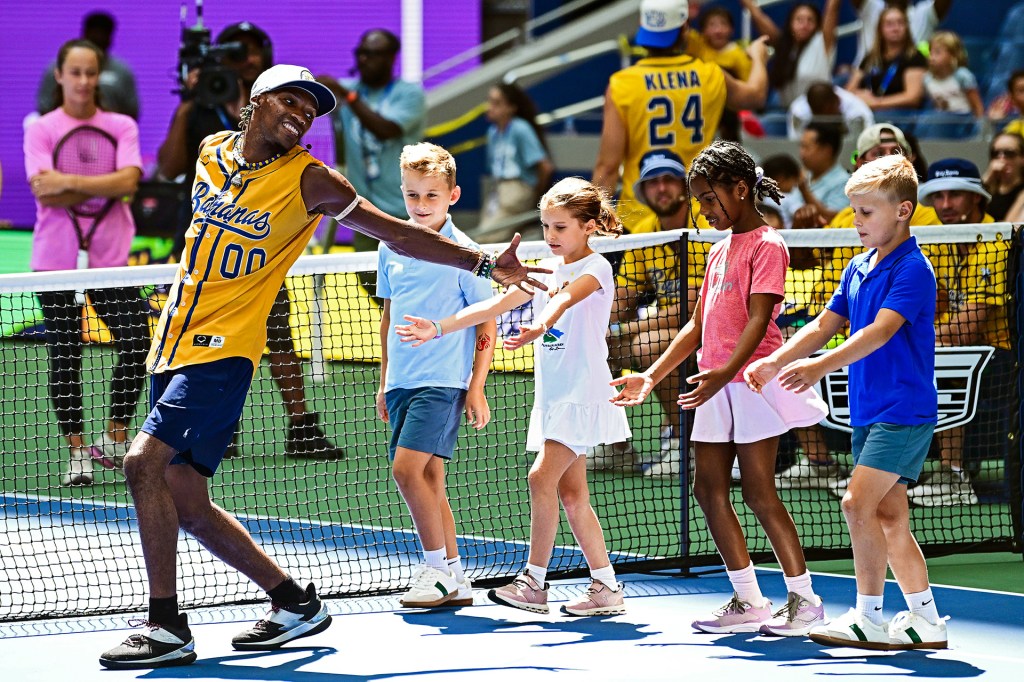 A man in a yellow sports jersey leads four kids walking in a line on a tennis court.