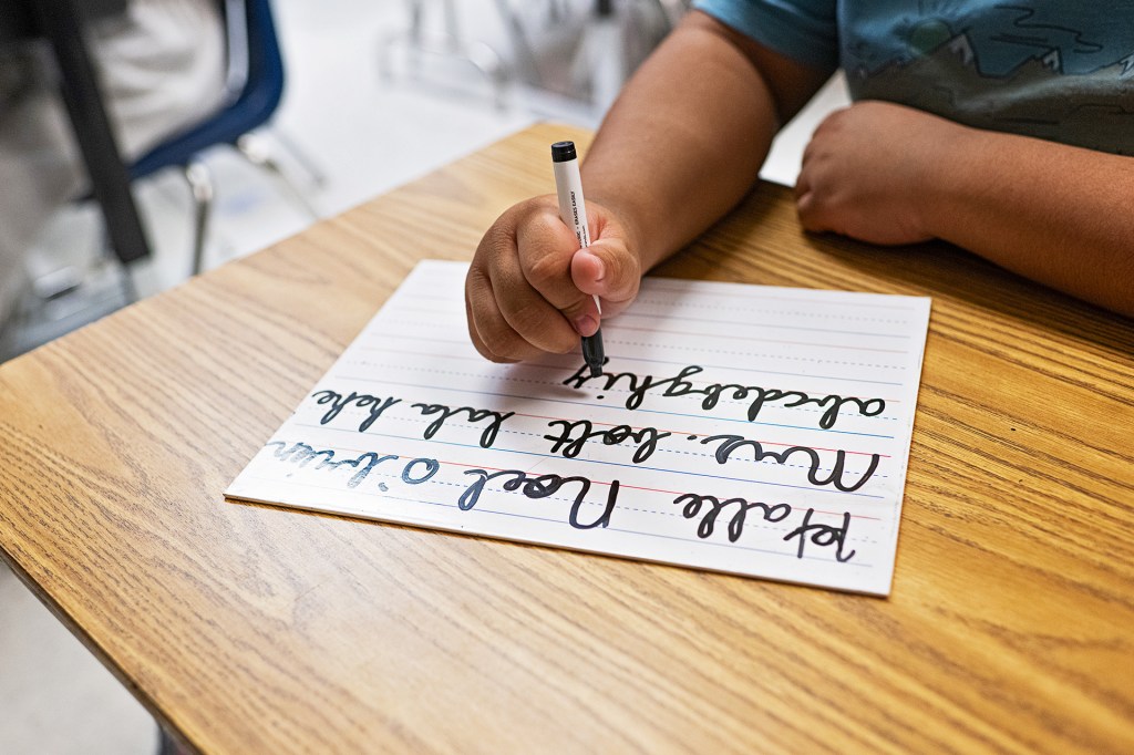 A student practices writing cursive letters with a marker at a classroom desk.