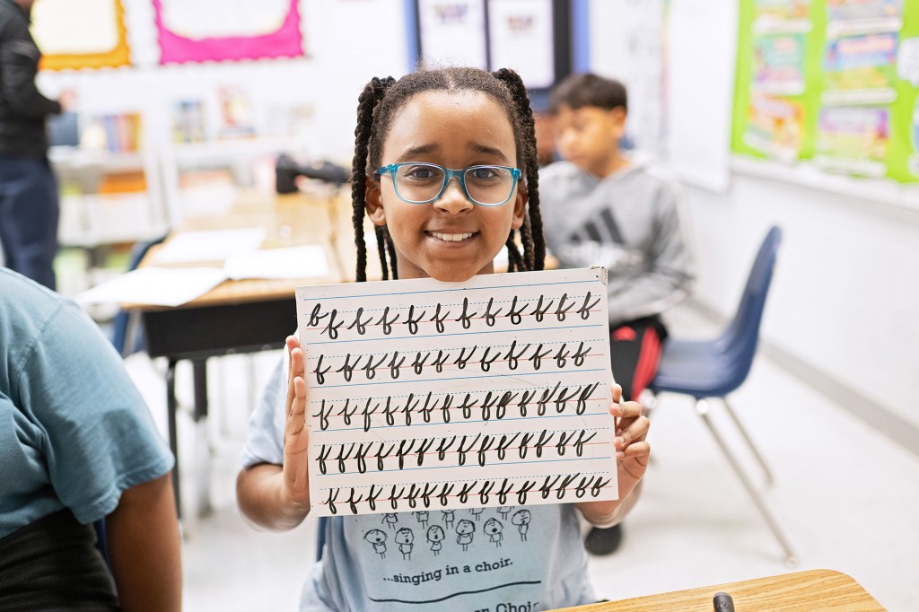 A smiling student holds up a board covered with rows of handwritten cursive letters.
