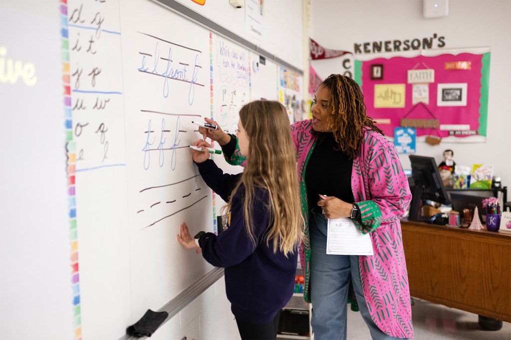A teacher helps a student practice writing cursive letters on a classroom whiteboard.