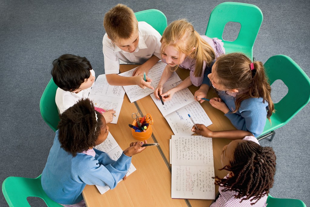 A group of children sitting at a table on green chairs working together.
