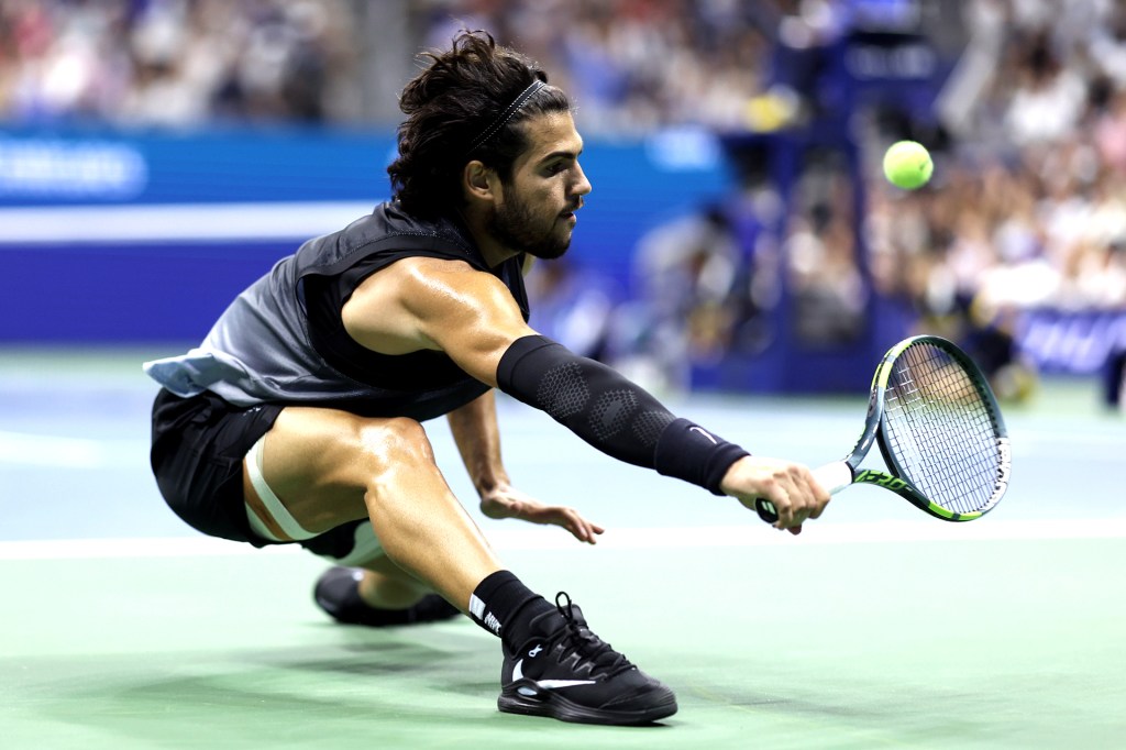 A tennis player wearing black in a deep lunge reaching for a ball.