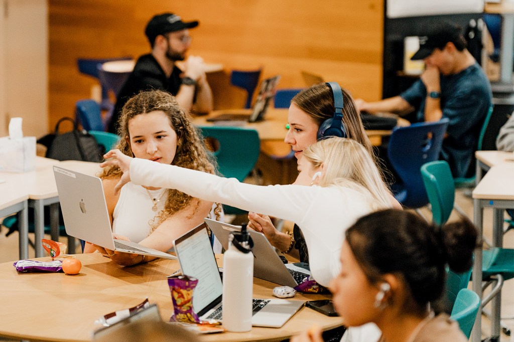 Students work together on laptops around a table in a classroom.