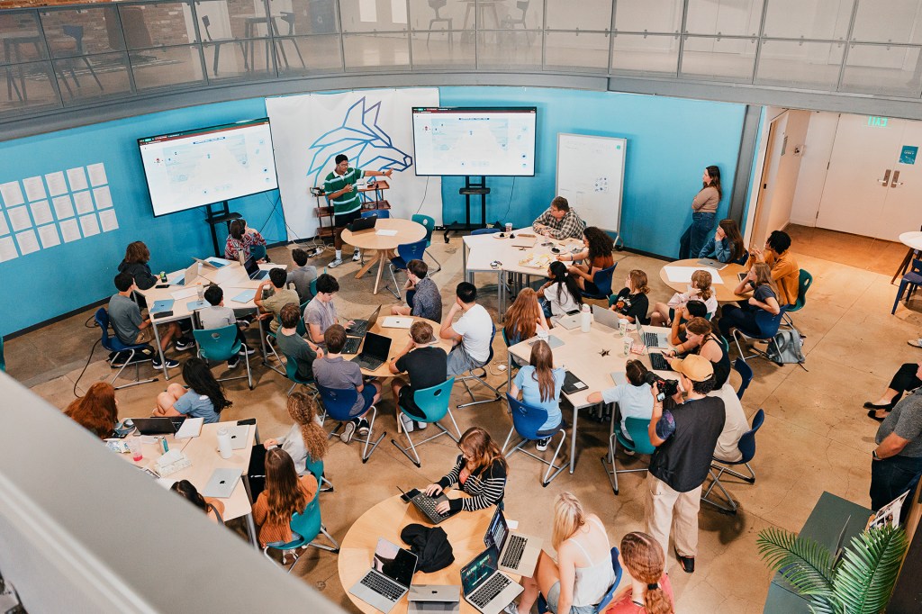 A large group of students attends a lesson in a classroom filled with tables and laptops.