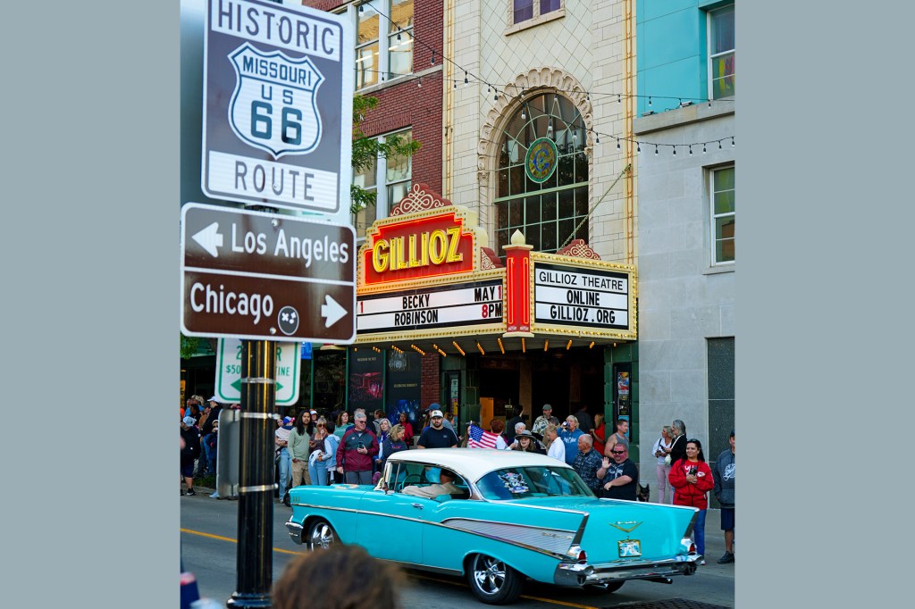 A vintage turquoise car drives past a theater and a sign marking a historic highway.