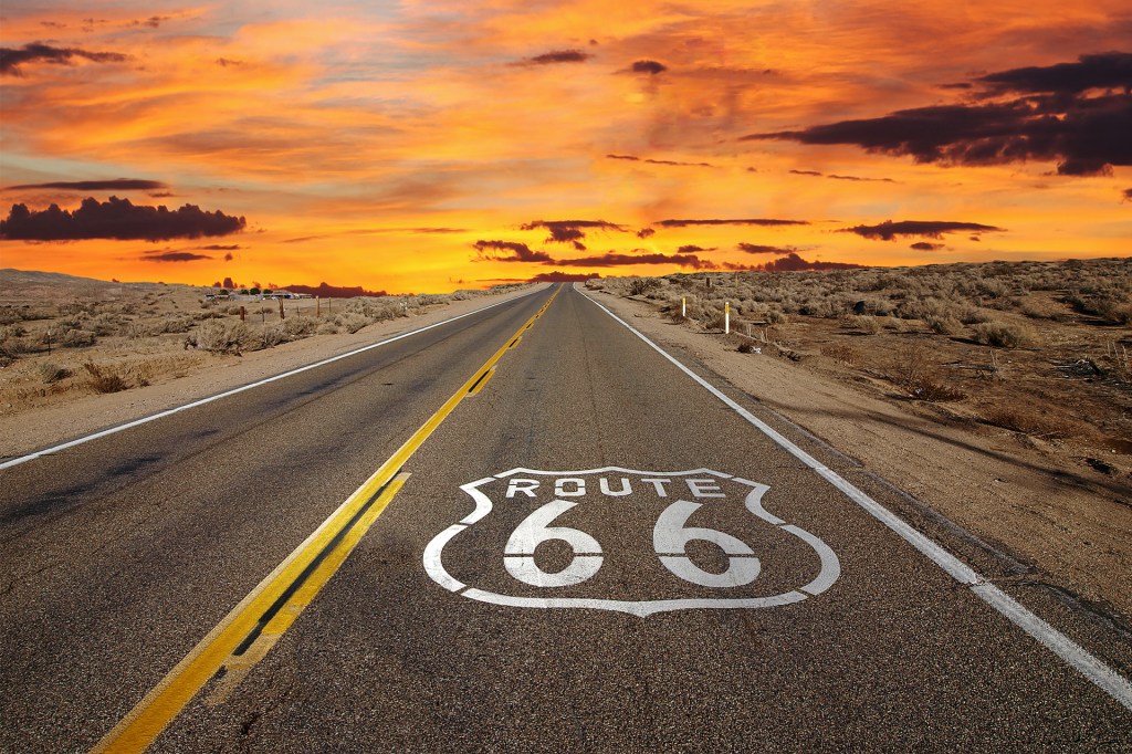 A highway marked with a large route shield stretches toward the horizon beneath a fiery sunset.