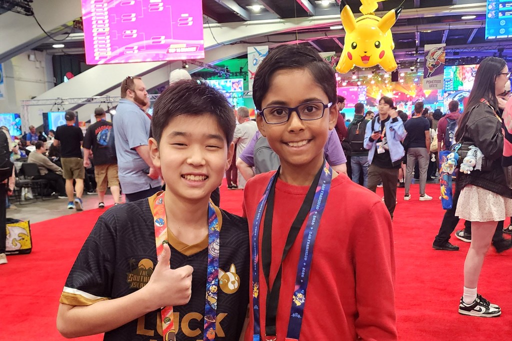 Two smiling children pose together at a crowded gaming event, with a large yellow character hanging overhead.