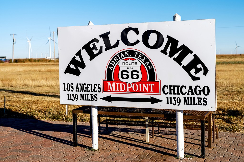 A roadside sign marks the halfway point between two major cities, with wind turbines in the distance.