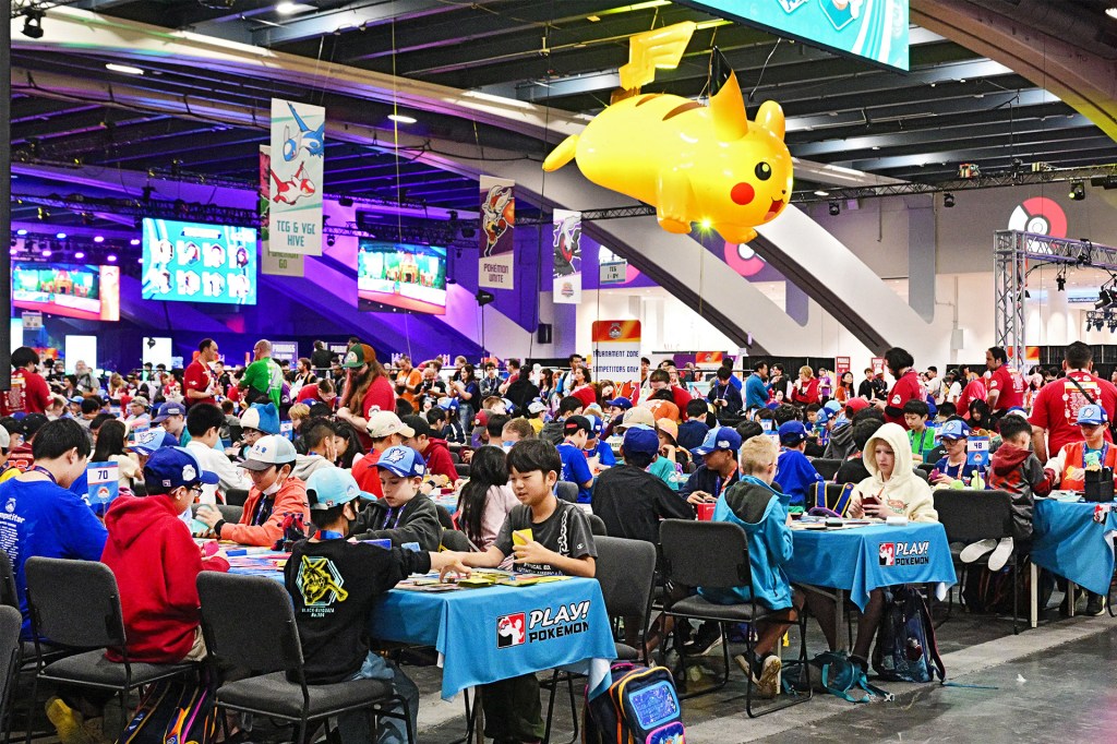 A crowded tournament hall is filled with people playing a trading-card game beneath a giant yellow character.
