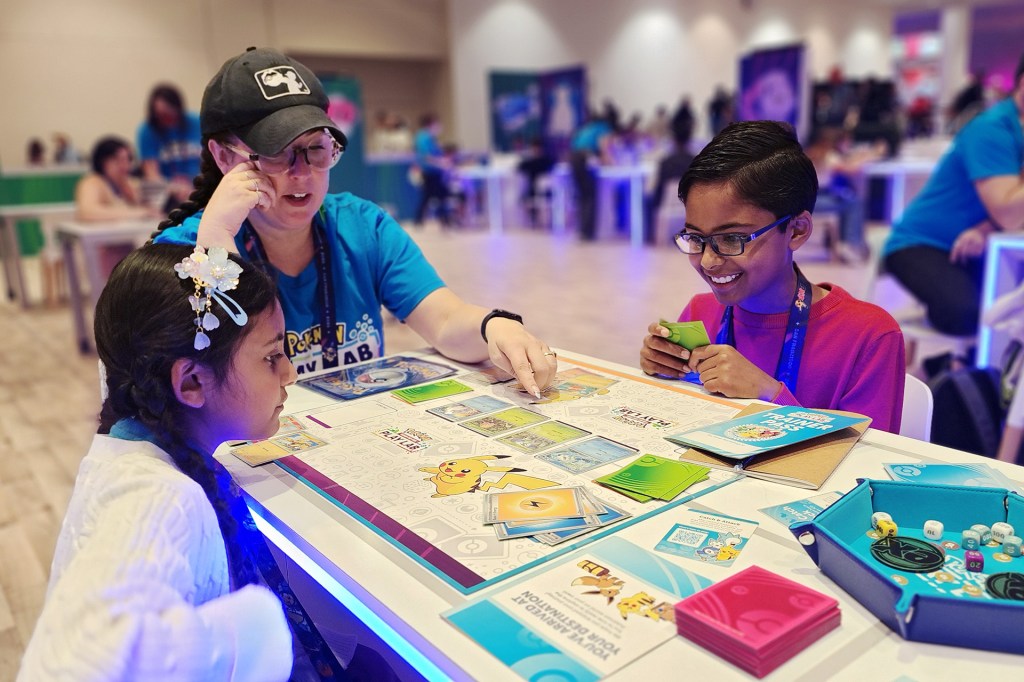 Two children play a trading-card game at a table while an adult helps them.