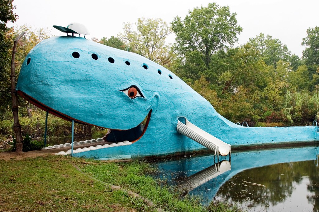 A giant blue whale-shaped roadside attraction sits beside a pond.