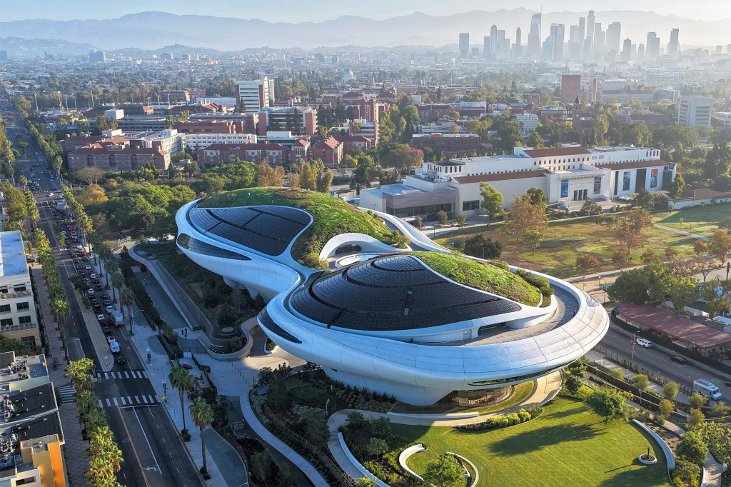 An aerial view shows a futuristic building with sweeping curves and green roofs, with a city skyline in the distance.