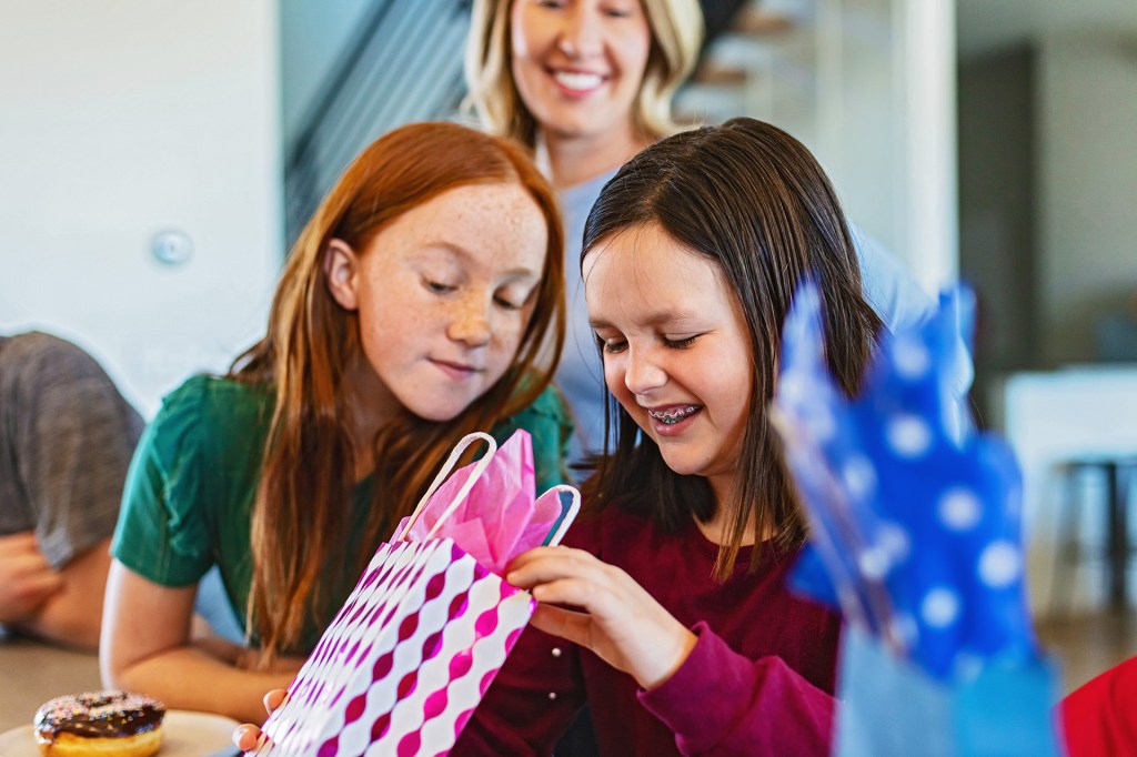 Two girls look inside a colorful gift bag while an adult stands behind them.