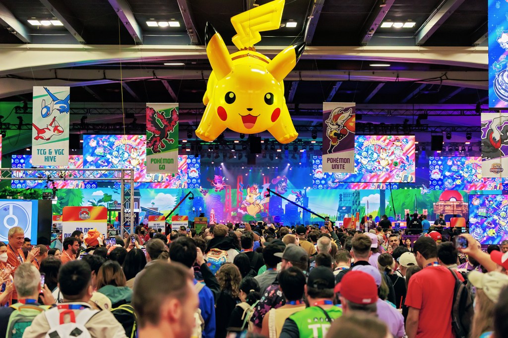 A large yellow cartoon creature hangs above a crowd at a busy gaming convention, with colorful screens and banners in the background.