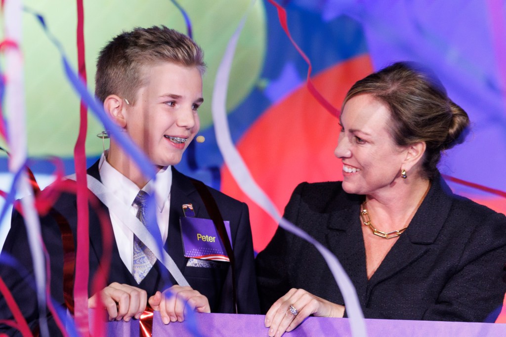A smiling boy in a suit and tie looks toward a woman onstage as colorful streamers hang around them during a celebration.