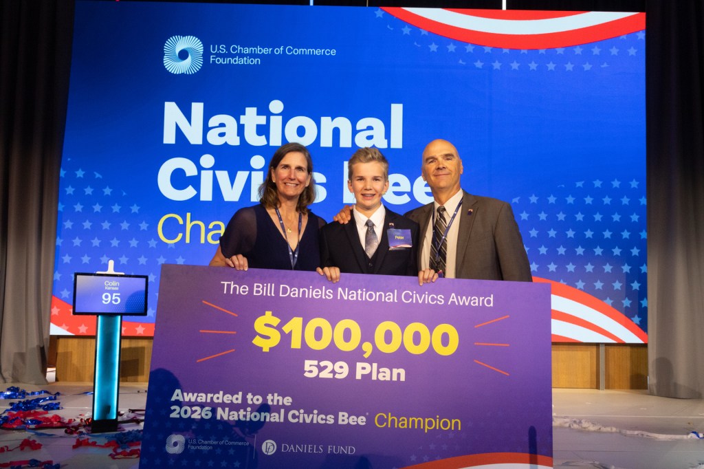 A boy in a suit stands between two smiling adults onstage, holding a large sign that says, "The Bill Daniels National Civics Award", with "$100,000" and "529 Plan" below it.