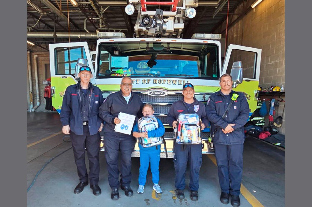 A boy holding a packed bag poses with firefighters in front of a fire truck.