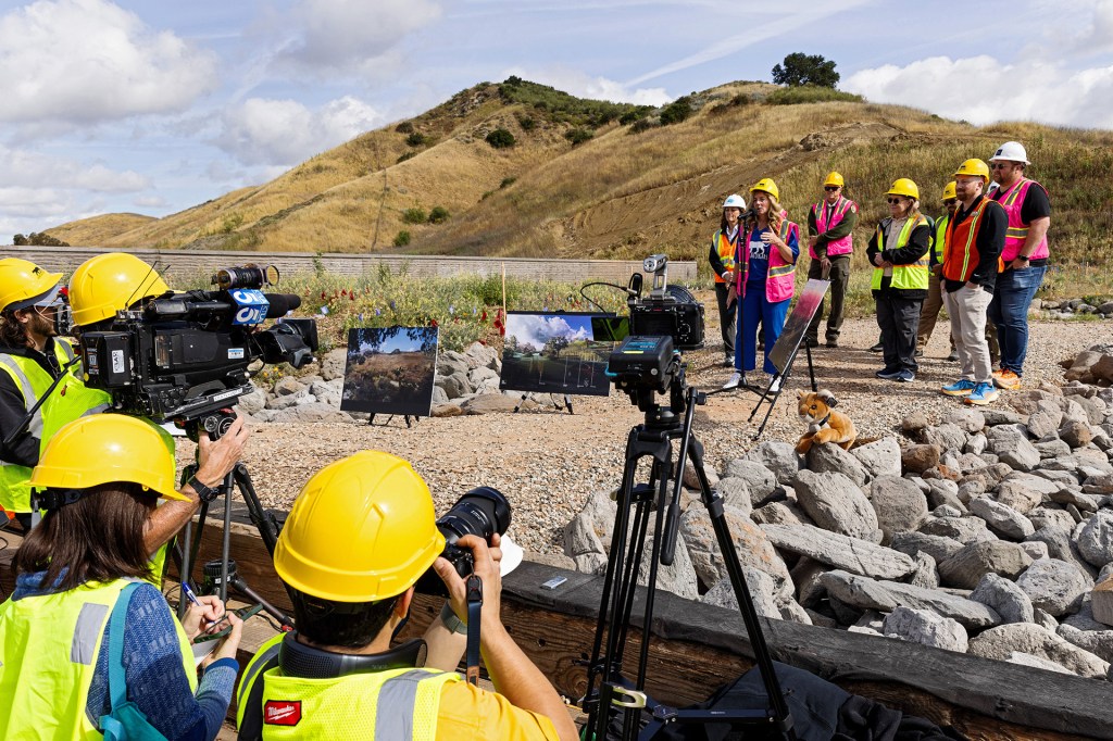 A woman in a hard hat and orange safety vest speaks into a microphone at an outdoor event to reporters and photographers in hard hats and safety vests.