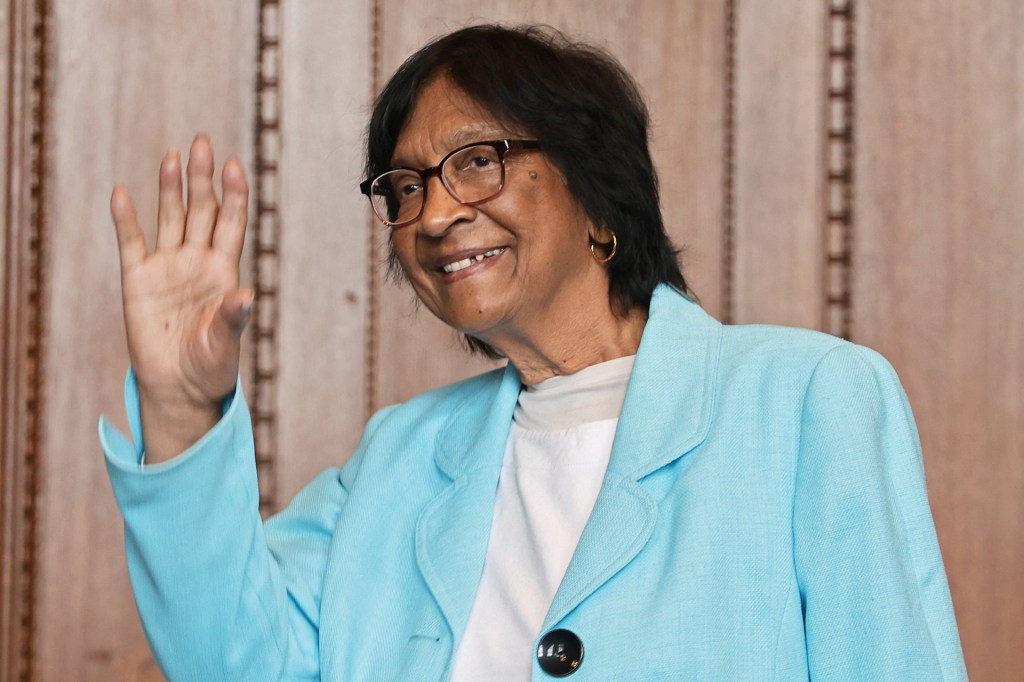 A woman wearing glasses and a light-blue jacket smiles and waves.