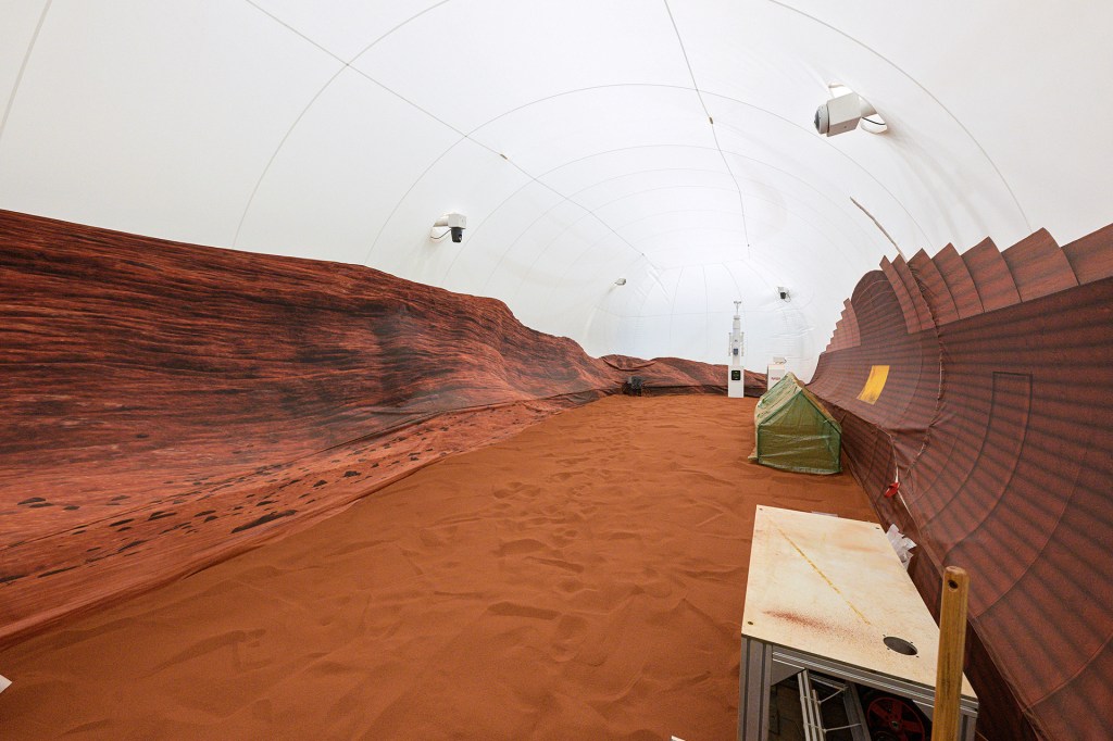 A large indoor habitat features red sand, rocky walls, a small green tent, and a curved white ceiling.