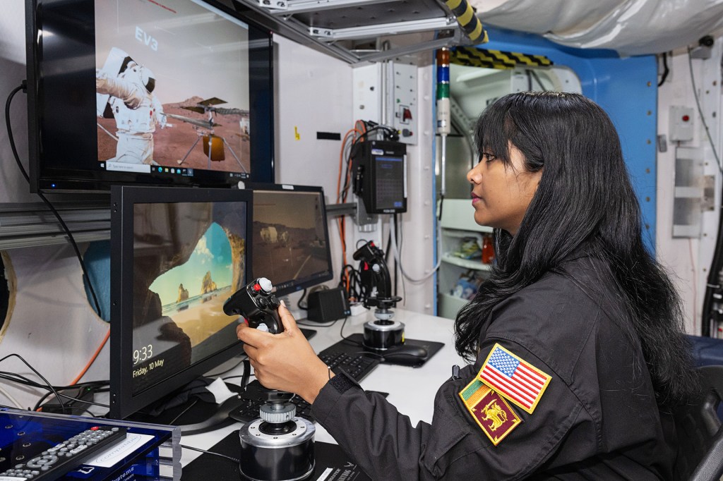 A woman wearing a black jumpsuit with a flag patch operates a joystick in front of computer monitors displaying a simulated space environment.