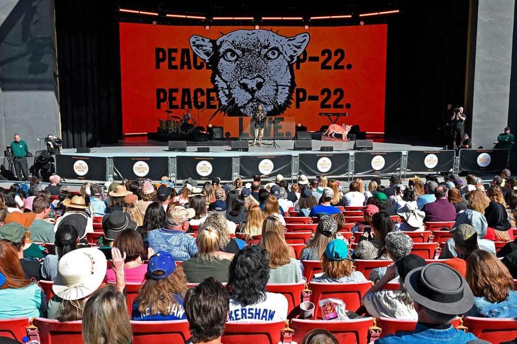A speaker stands onstage before a large screen featuring a mountain lion's face, addressing an audience seated outdoors.