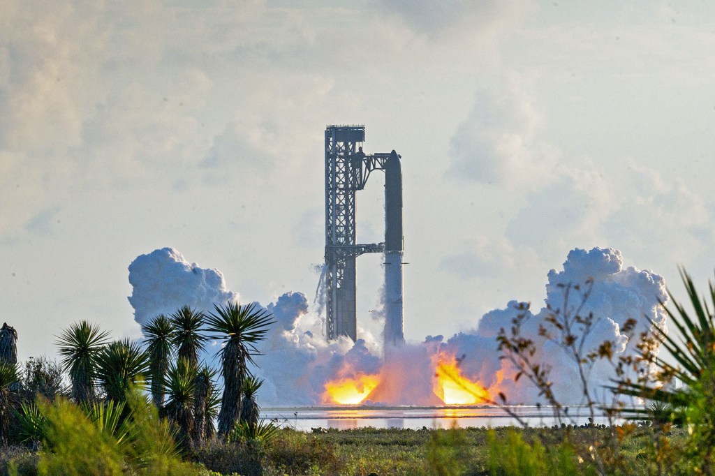 A large rocket stands on a launchpad as flames and thick clouds of smoke rise from its base.