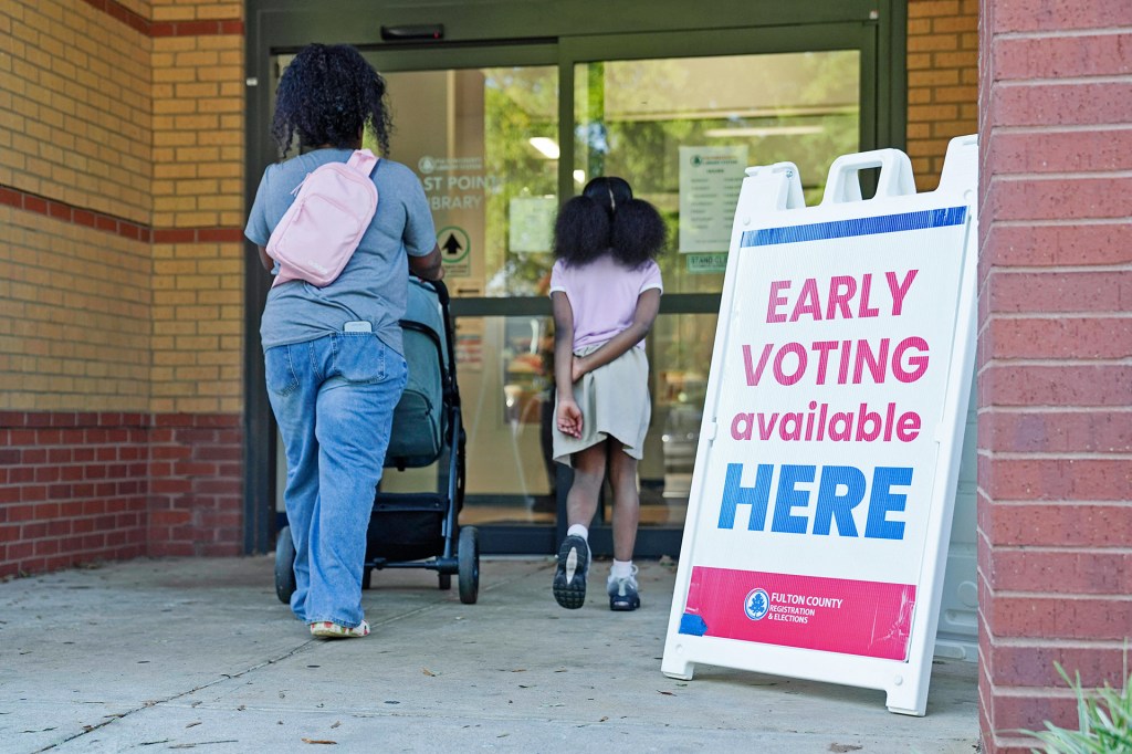 A woman pushes a stroller toward a building entrance beside a girl and a sign announcing early voting.