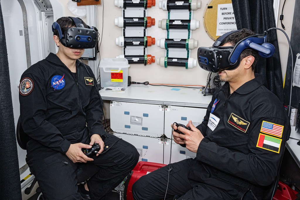 Two men wearing black jumpsuits and virtual-reality headsets sit facing each other while holding controllers inside a training facility.